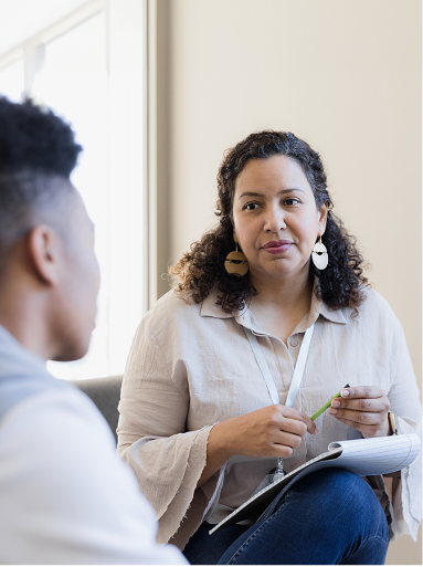 Woman holding a clip board and pen listens to a young person seated across from her.